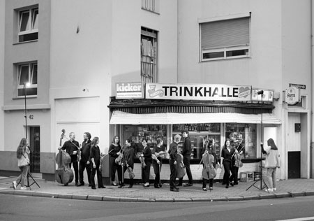 Schwarz-weiß Foto vom Ensemble Ruhr: einige Menschen mit Streichinstrumenten stehen beieinander an einer Trinkhalle, Foto: Jan Pauls