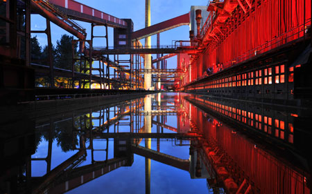 Blick auf Zeche Zollverein in Essen, auf der rechten Seite leuchtet sie rot, im blauen Wasser unten spiegelt sich der obere Teil des Bildes, Foto: Ruhr Tourismus/Vinken