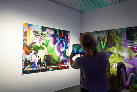 Die Uzwei-Leiterin Mirjam Gaffran scannt mit einem Tablet das Gemälde „Schwebende Malerei“ von Eunjeong Kim ab und auf dem Bildschirm des Tablets bewegen sich die farbenfrohen Elemente des Ölgemäldes, Foto: Miu-Wah Lok